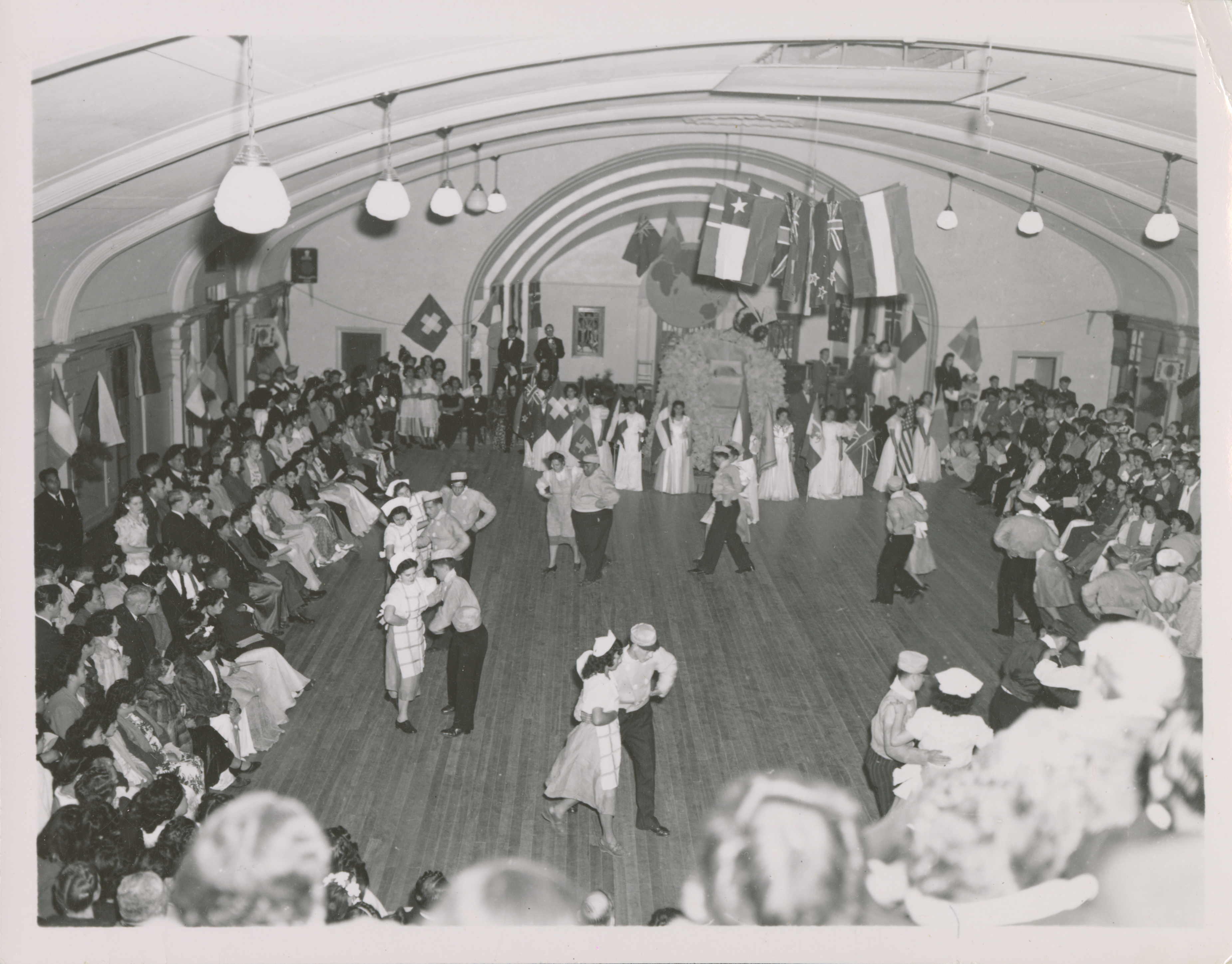 <strong>Gold and Green Ball, New Zealand.</strong> Circa 1949. Young women and young men in New Zealand sashayed around the dance hall as they performed the floor show, observed by dozens of spectators and the ball royalty. These elaborate, formal balls became a cherished annual tradition of Mutual Improvement Associations beyond North America and often involved local church members and non–Latter-day Saints alike, including individuals outside the traditional MIA age. (Image courtesy Gordon Woodruff Young, copy at Church History Library, Salt Lake City.)