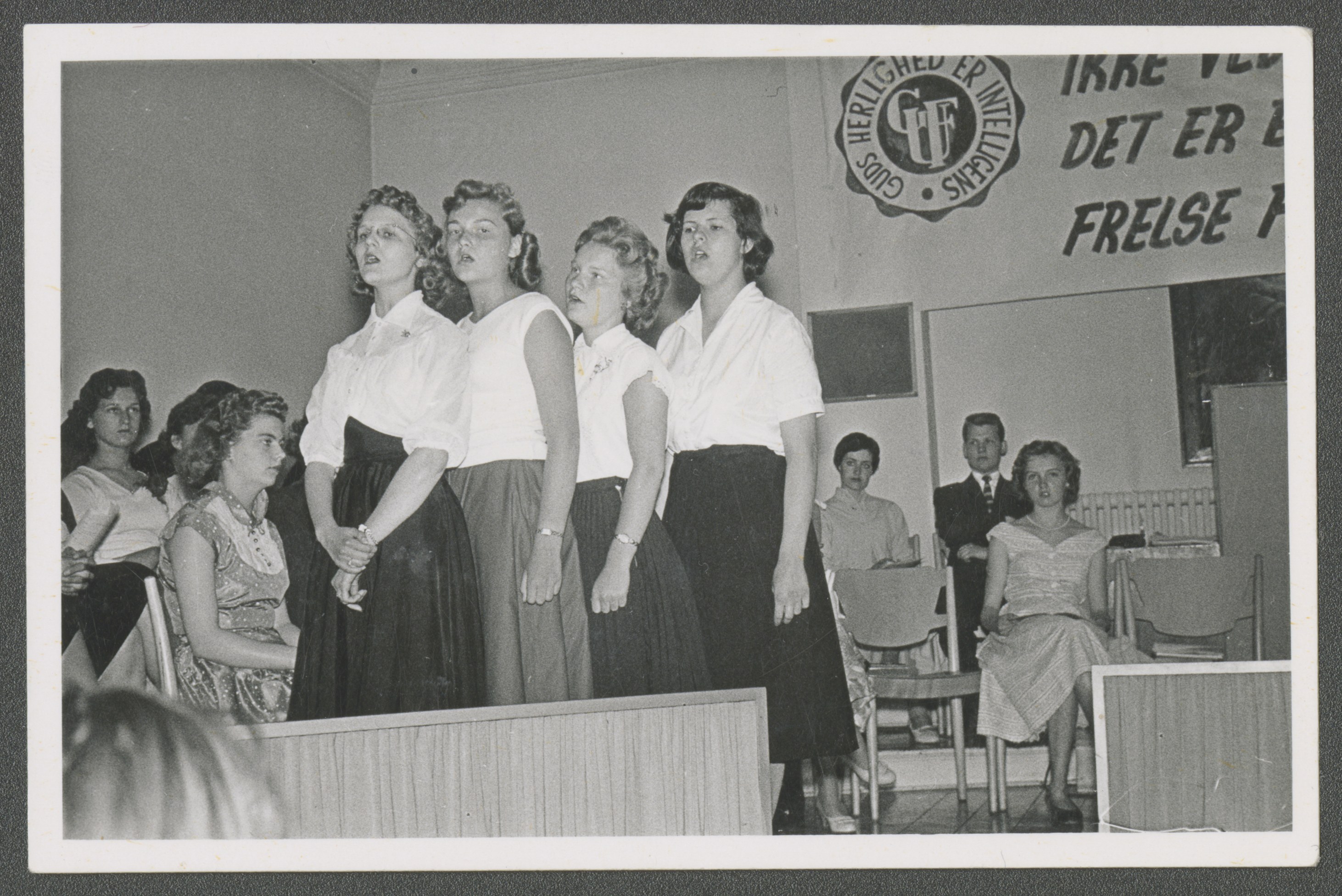 <strong>Singers at the Danish Mission MIA convention.</strong> 1956. Young women from Copenhagen, Denmark, participated in a music contest in Aalborg, Denmark. A banner displaying the year’s MIA theme is in the background: “I am not ashamed of the gospel of Christ: for it is the power of God unto salvation to every one that believeth” (Romans 1:16). Similar regional conventions were held all over the world. (Image courtesy Church History Library, Salt Lake City.)